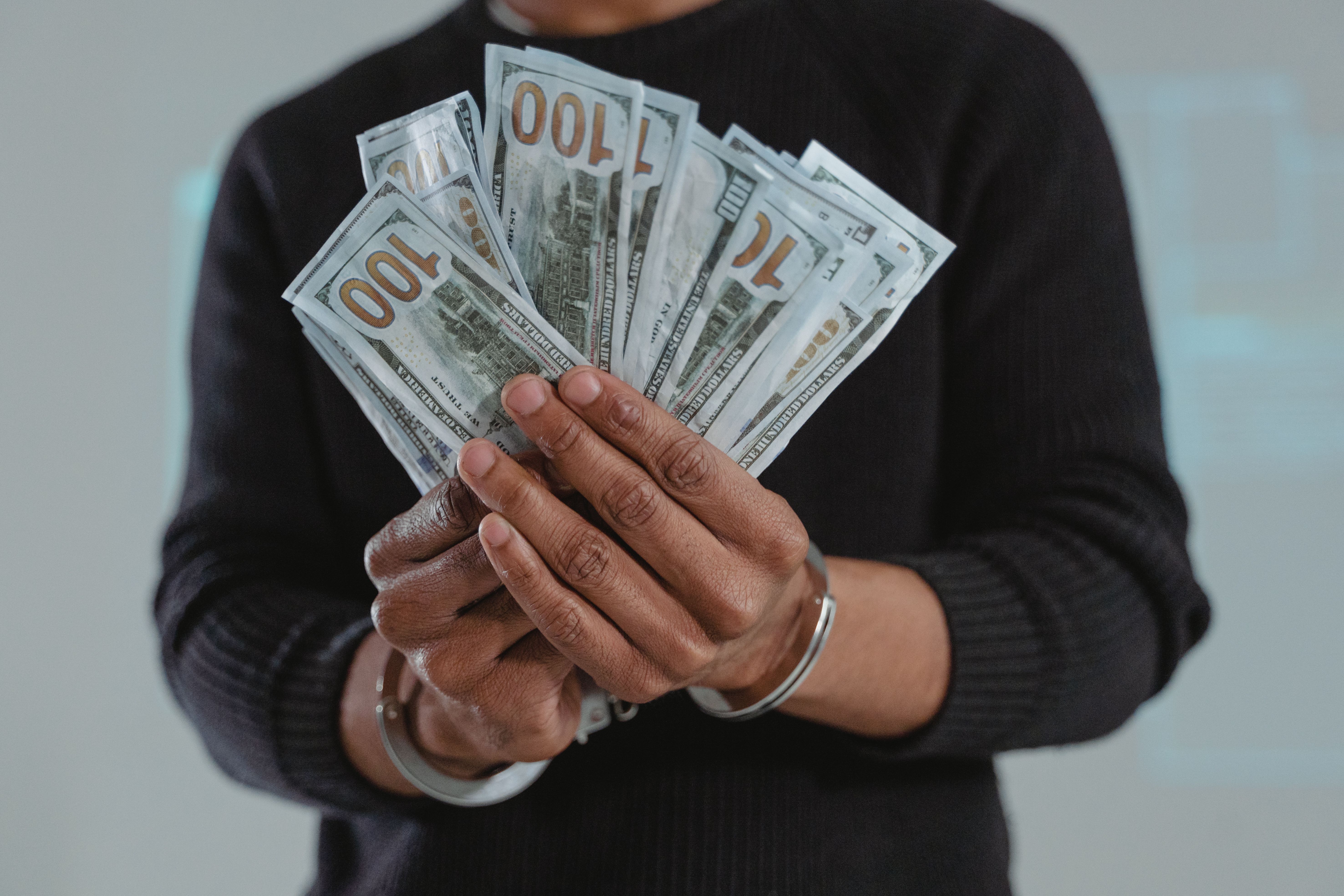 Handcuffed hands holding 100-dollar bills, symbolizing greed, corruption, and personal gain
